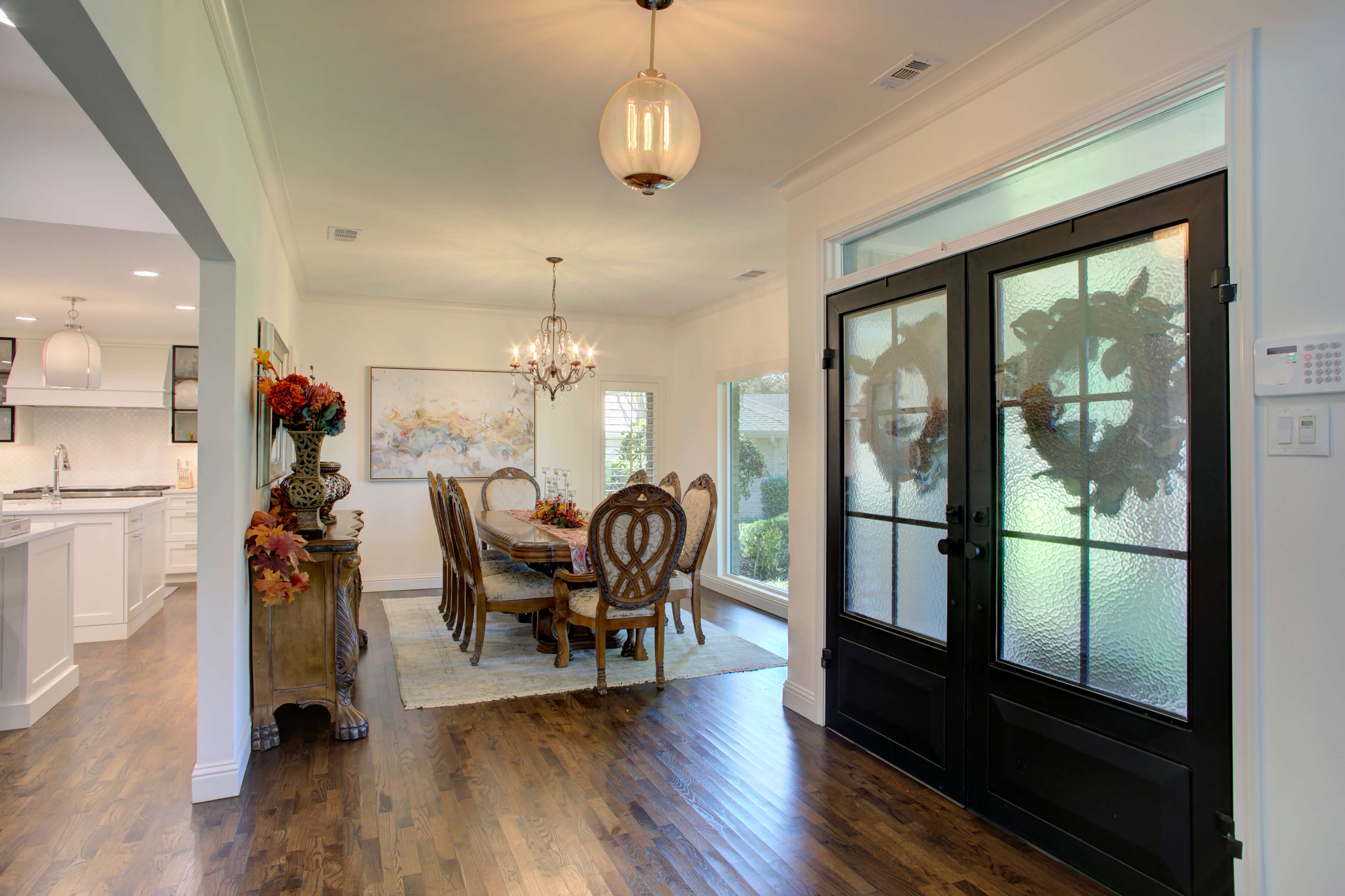 A dining room with a long table and chairs, large windows, and a chandelier. Double doors with glass panels lead to the room, and a view of a connected white kitchen is visible.