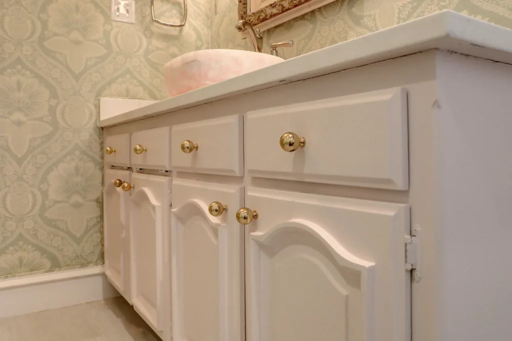 Close-up view of a bathroom vanity with white cabinet doors and gold knobs, featuring a round vessel sink and patterned wallpaper in the background.
