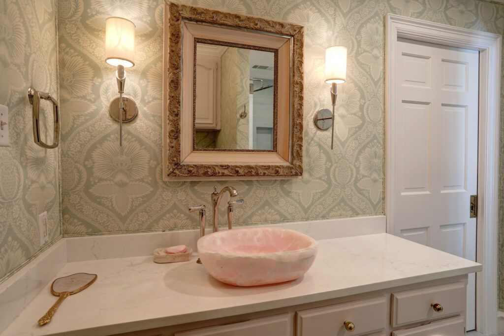A bathroom vanity with a pink marble vessel sink, ornate framed mirror, wall sconces, and patterned wallpaper. A hand mirror and soap tray are on the white countertop.