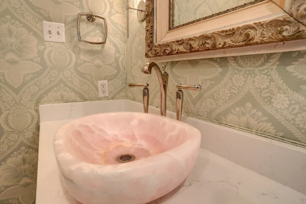 A pink stone vessel sink with a gold faucet sits on a white countertop in a bathroom with patterned wallpaper and a decorative mirror.