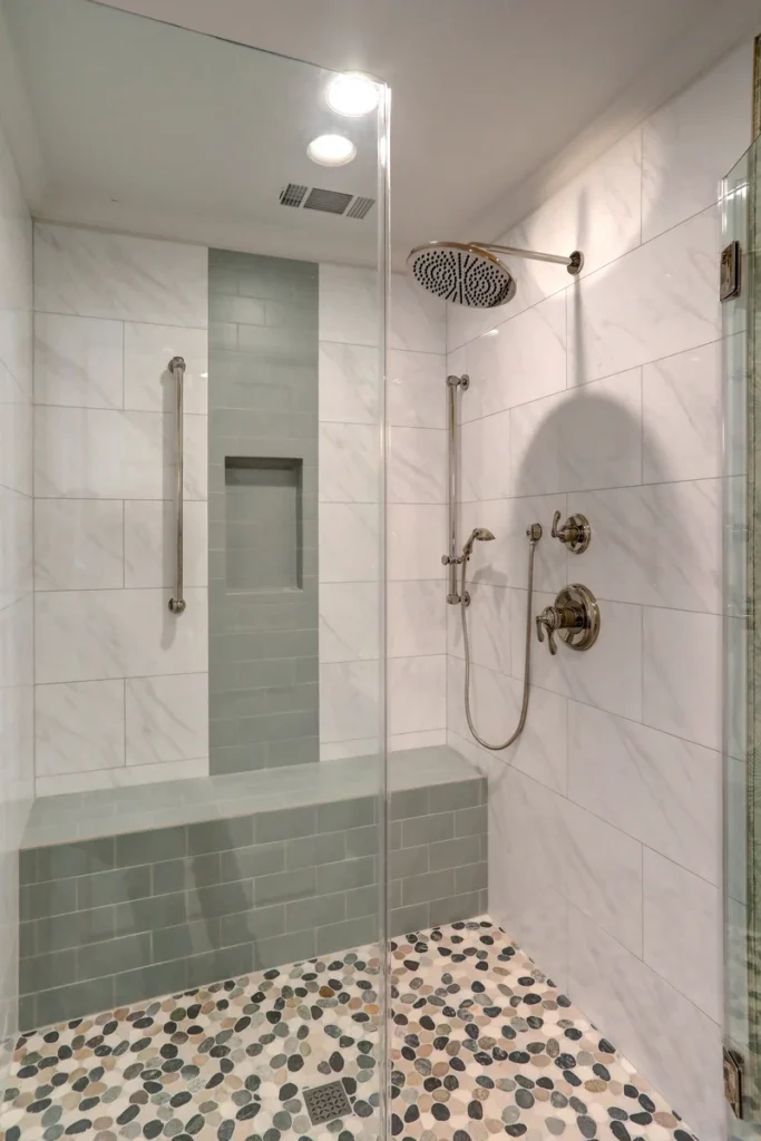 Modern walk-in shower with a built-in bench, wall niche, grab bar, rainfall showerhead, handheld shower, and pebble tile floor.