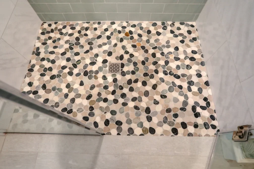 A shower floor with pebble-style tiles in various colors, a central square drain, and glass door partially visible in the foreground.