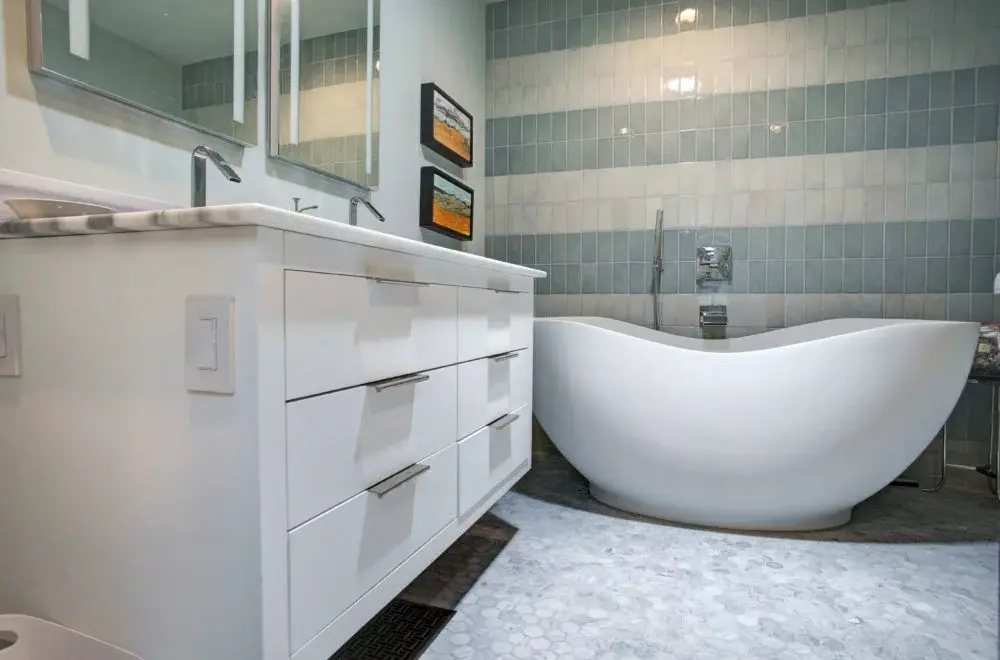 Modern bathroom with a white freestanding bathtub, white vanity with drawers, double sinks, and striped tiled wall in neutral tones.