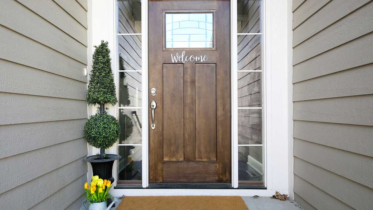 Dark Wooded Door Leading to House with Welcome Sign on Front A wooden front door with glass panels, a welcome sign, potted topiary and yellow tulips, and a doormat on a concrete porch.