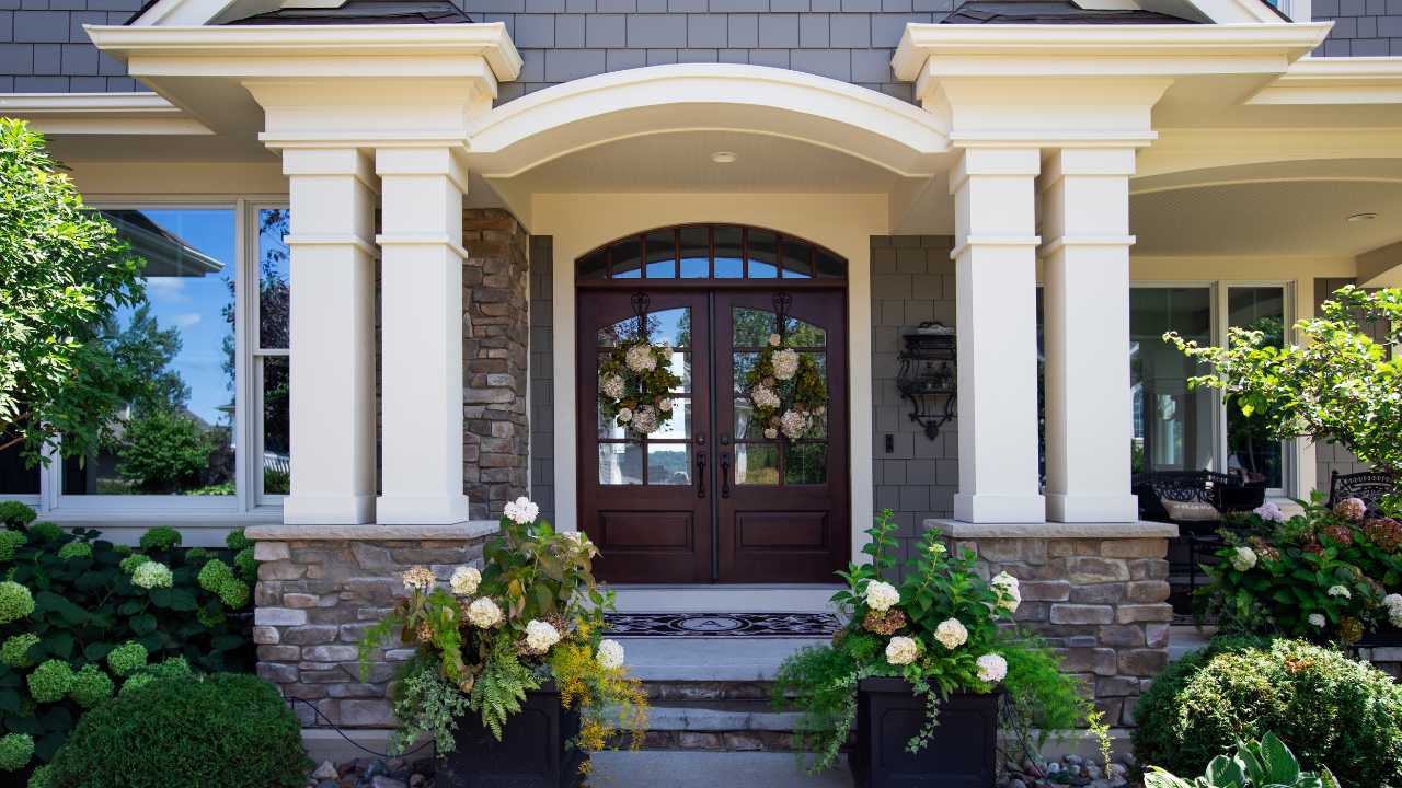 Dark Wooded Door Leading to House with Wreaths On Top and White Archway Front entrance of a house with double wooden doors, stone and siding exterior, white columns, and flower planters on either side of the steps.