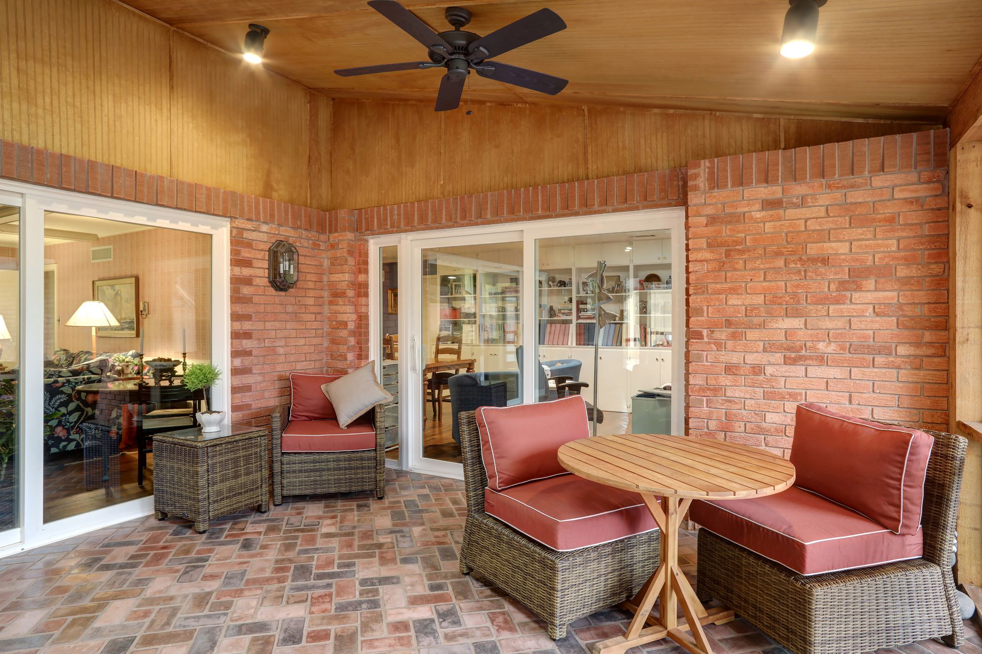 Enclosed patio with brick walls and floor, wicker furniture with red cushions, a round wooden table, ceiling fan, and sliding glass doors leading to interior rooms.