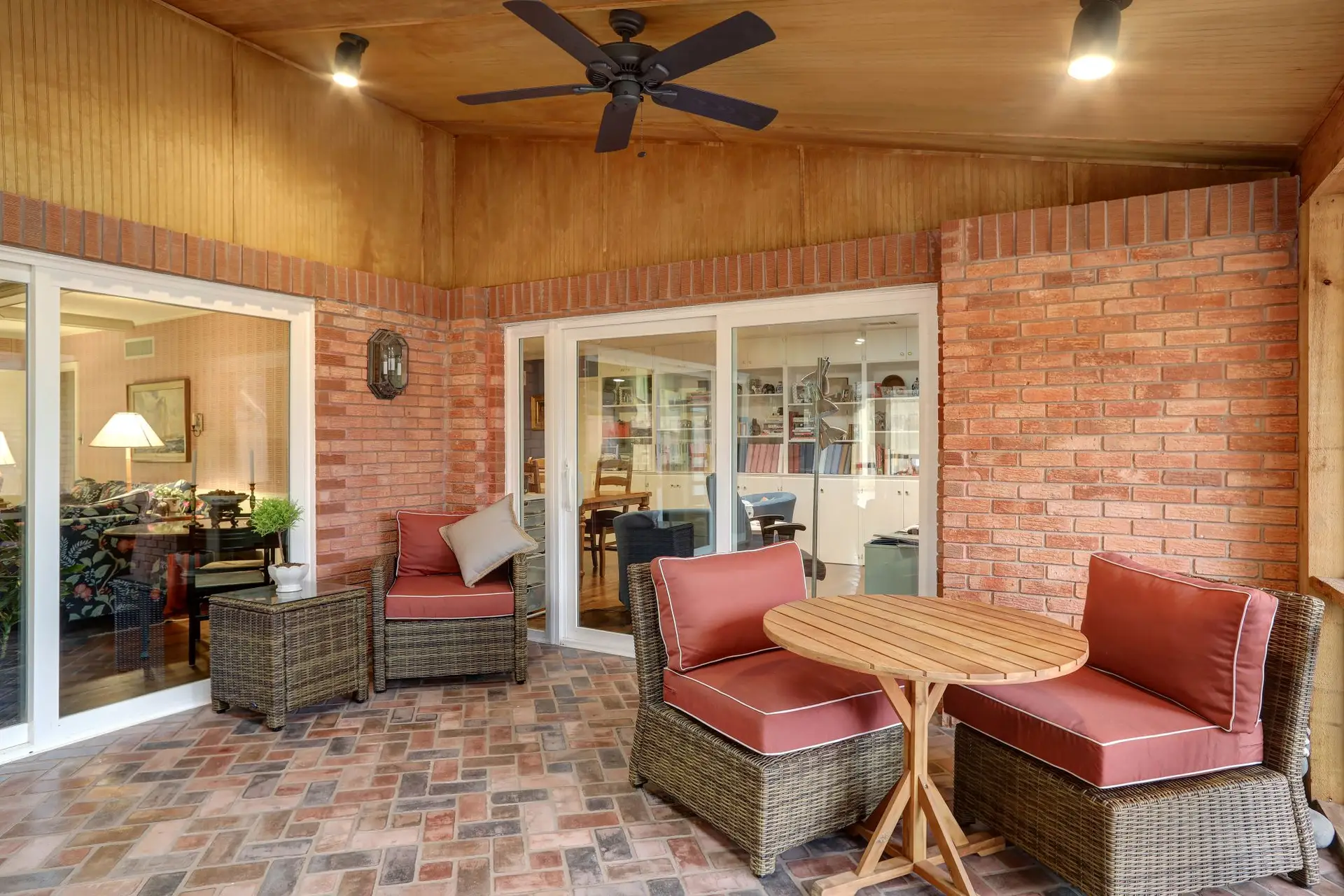 Enclosed patio with brick walls, sliding glass doors, wicker furniture with red cushions, a round wooden table, ceiling fan, and overhead lighting.
