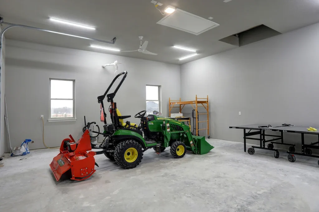 A green utility tractor with a red attachment is parked in a spacious garage with gray walls; a ping pong table and a yellow ladder are visible in the background.
