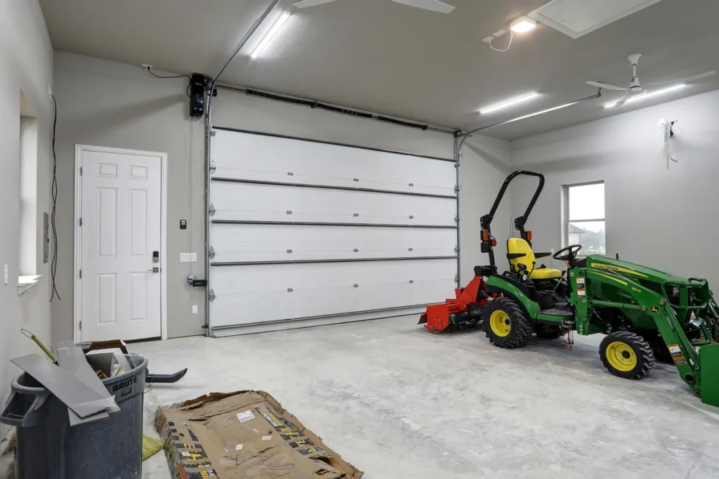 A clean garage interior with a closed white garage door, a side entry door, two green tractors, and trash bins on a bare concrete floor.