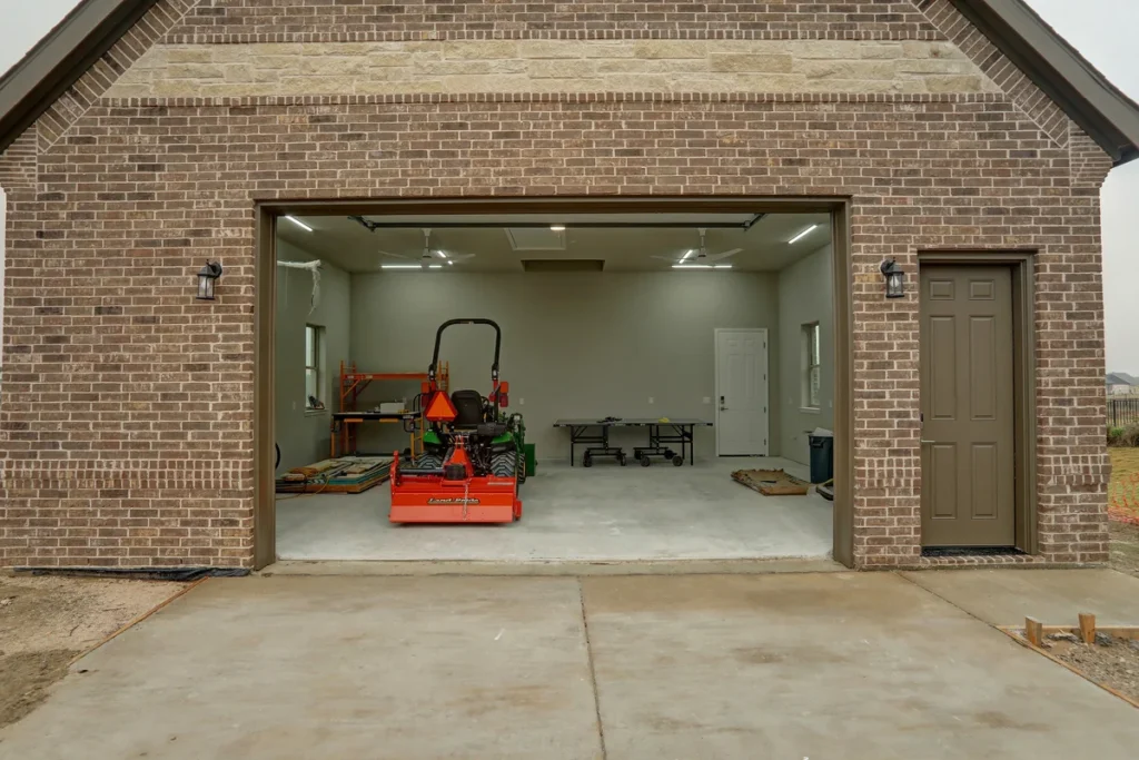 A brick garage with an open door shows a compact tractor with attachments, a folding table against the back wall, and various items along the sides on a concrete floor.