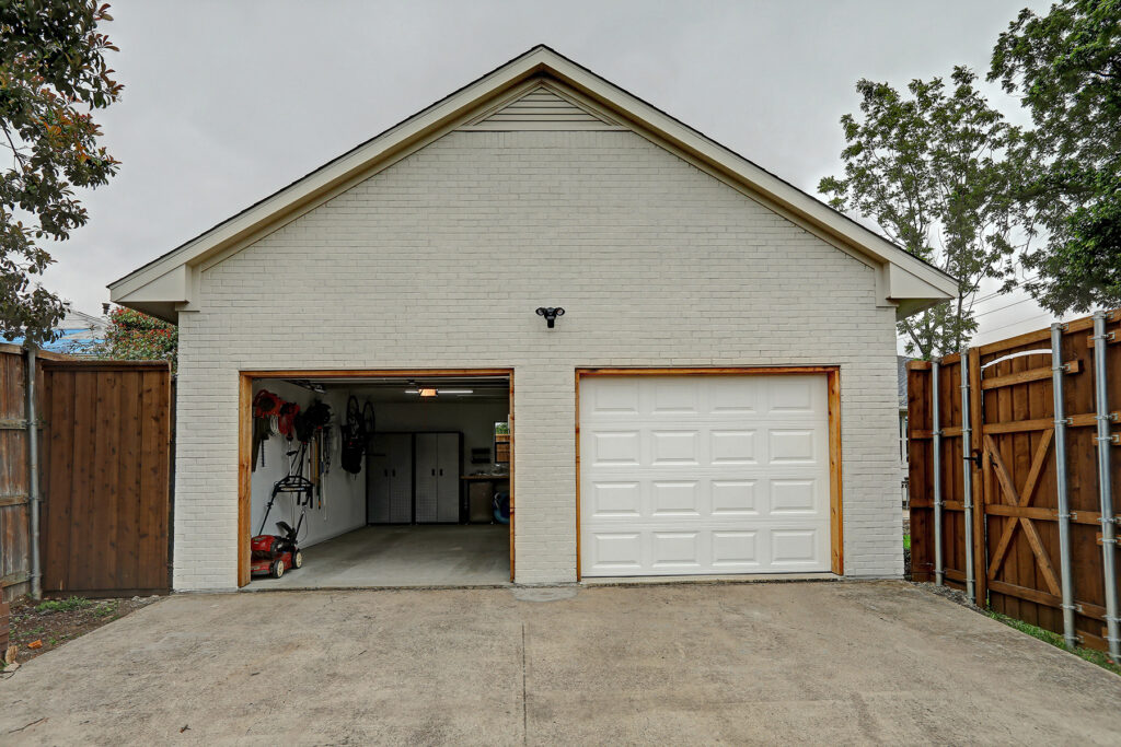 A detached two-car garage with one door open, showing an empty interior and tools mounted on the wall, set in a fenced backyard.