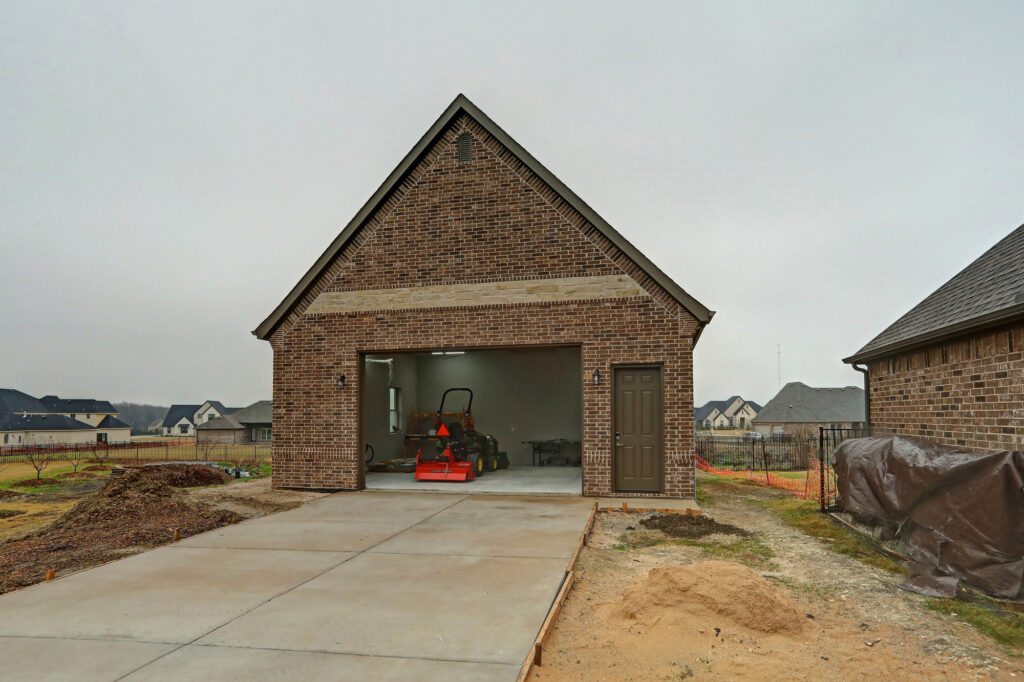 A newly constructed brick house with an open garage door, a driveway, construction equipment inside, and materials scattered around the yard.