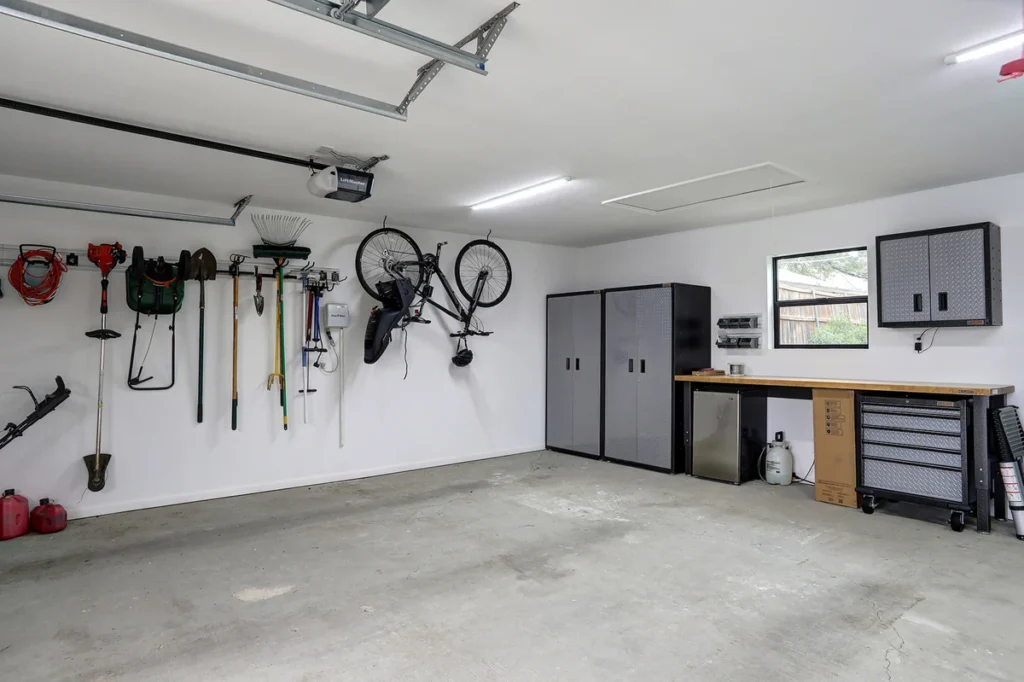 Organized garage with tools and garden equipment hanging on the wall, two bicycles mounted, storage cabinets, a workbench, and a window. The floor is clean and empty.