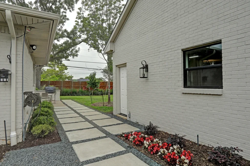 A concrete walkway with square pavers leads between a white brick house and garage, bordered by small plants and gravel.