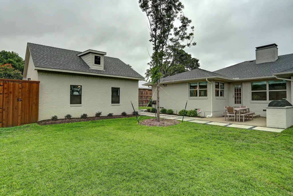 A backyard with green grass, a central small tree, a patio with outdoor furniture, and two light-colored brick buildings under a cloudy sky.