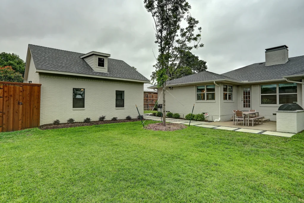 A fenced backyard with green grass, a tree, a detached garage, and a patio area with outdoor furniture next to a light-colored house.
