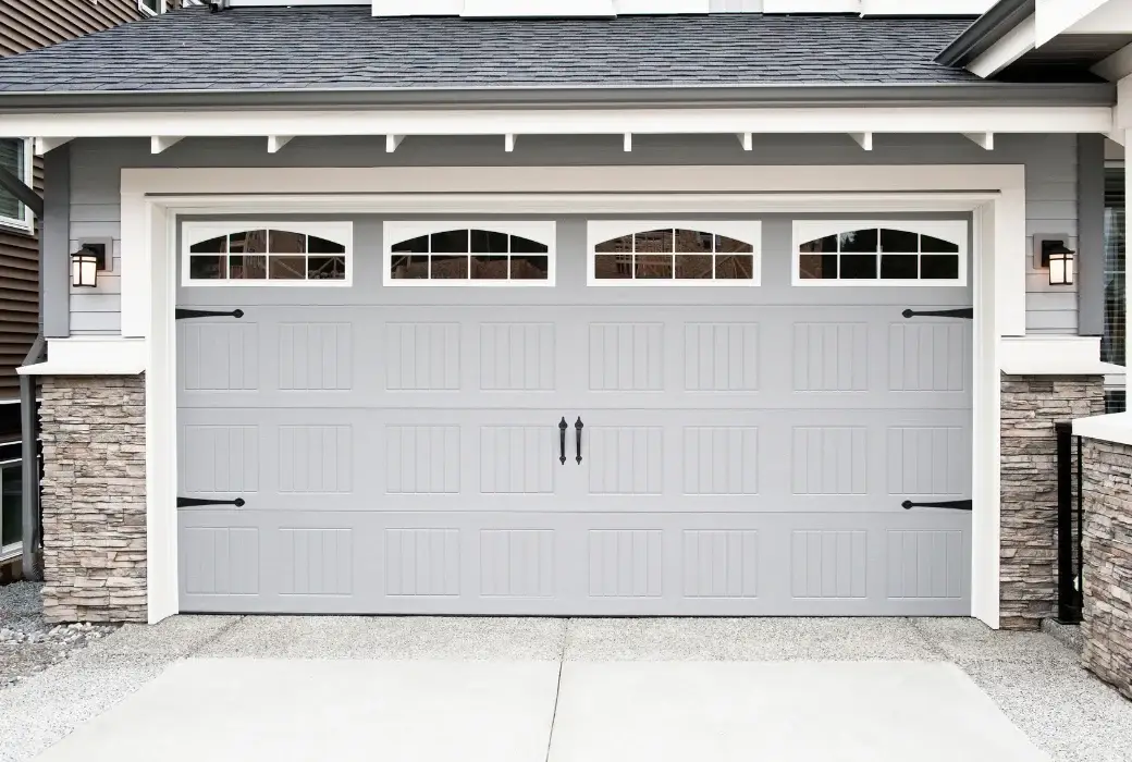 A closed light gray garage door with four small rectangular windows and decorative black handles, flanked by stone columns and exterior wall lights.