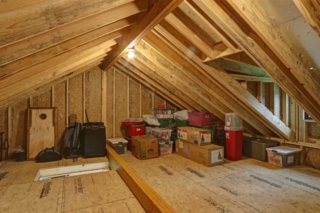 Unfinished attic with exposed wooden beams and insulation, illuminated by a single bulb, with boxes and storage containers arranged along the walls.