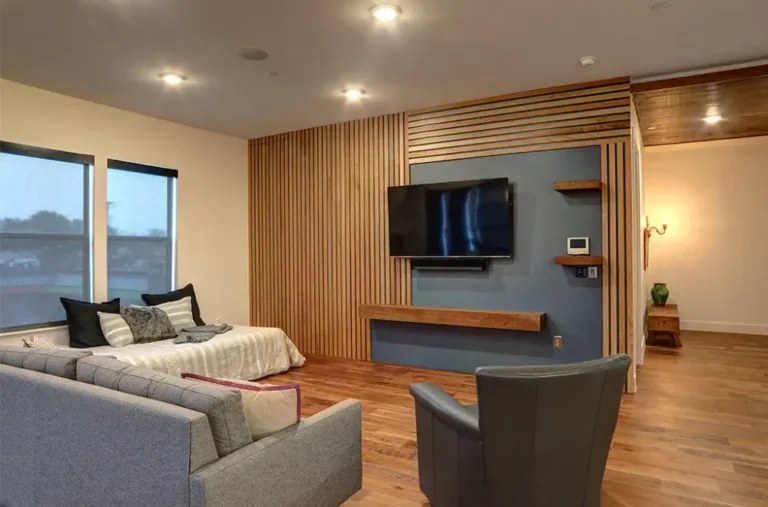 Modern living room with wood flooring, a gray armchair, a sectional sofa, a bed, and a wall-mounted TV on a blue accent wall with wooden slats.