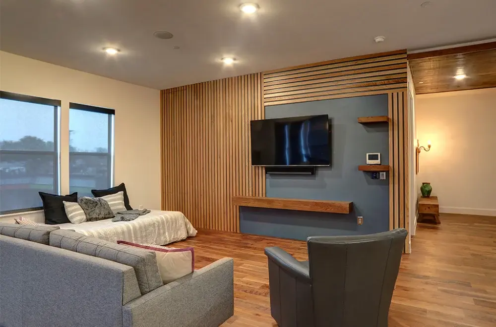 Modern living room with wood flooring, a gray armchair, a sectional sofa, a bed, and a wall-mounted TV on a blue accent wall with wooden slats.