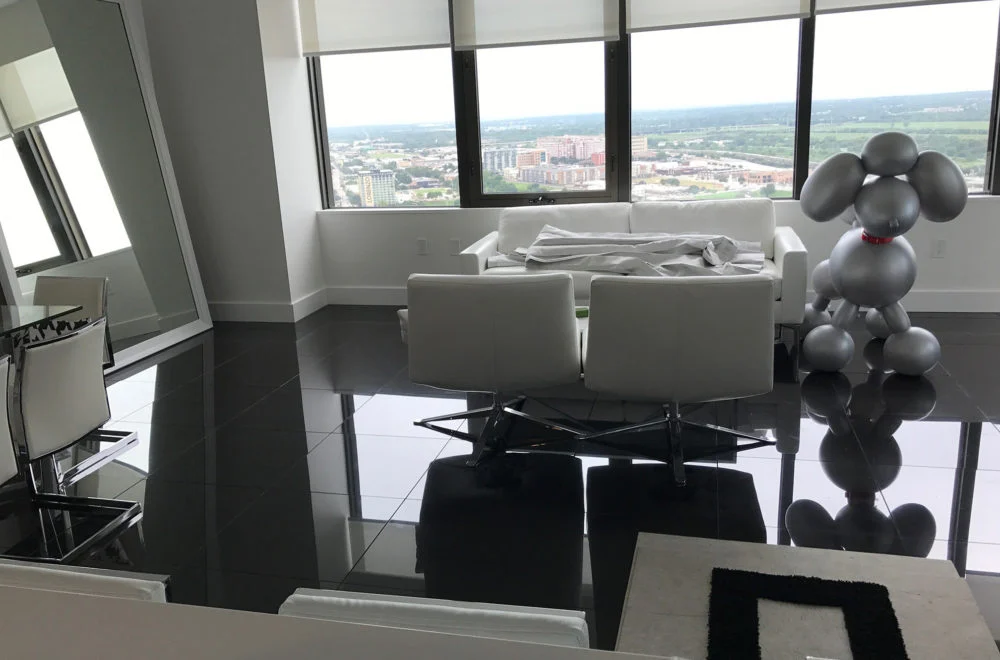 Modern living room with glossy black floor tiles, white furniture, large windows overlooking a city, and a silver balloon dog sculpture in the corner.