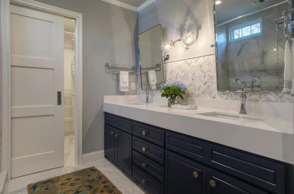 Modern bathroom with a double-sink vanity, navy blue cabinets, marble backsplash, and wall-mounted mirrors; a small bouquet of flowers sits on the counter.