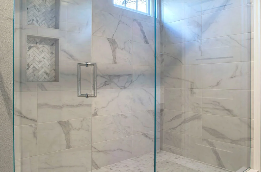 Modern walk-in shower with large marble tiles, glass door, built-in shelves with herringbone tile pattern, and a window near the ceiling.