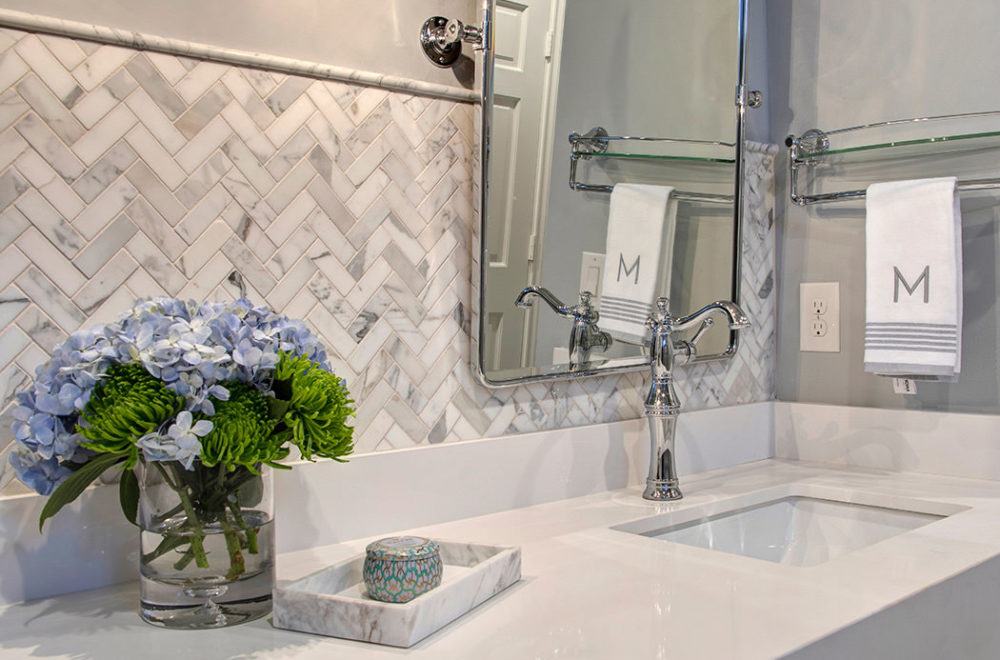 Modern bathroom vanity with a marble herringbone backsplash, glass vase of blue and green flowers, monogrammed towel, and a large mirror above the sink.