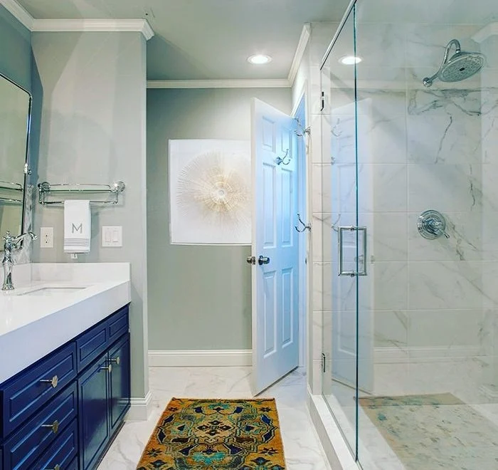 Modern bathroom with blue vanity, white countertop, glass shower with marble tiles, colorful rug, and wall art above a towel rack. The door is open, and lighting is recessed.