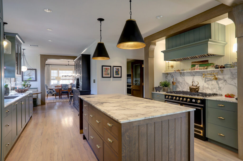 Modern kitchen with green cabinetry, marble countertops, a central island, brass hardware, pendant lights, and a gas stove set against a marble backsplash. Dining area visible in background.