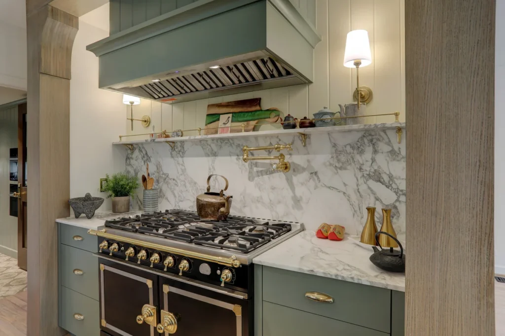 A modern kitchen features a black and gold stove, marble backsplash, open shelves with decor, and brass fixtures under a green range hood.