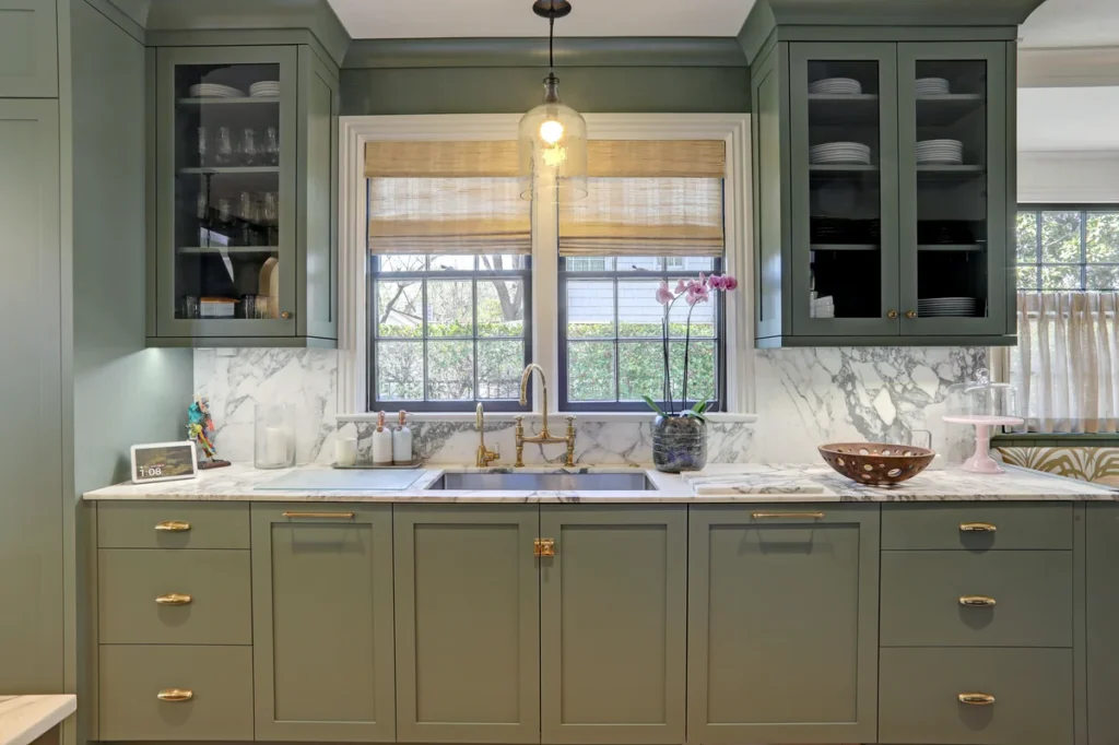 A modern kitchen with green cabinets, marble countertops and backsplash, glass-front upper cabinets, a sink, and a window with bamboo shades above.