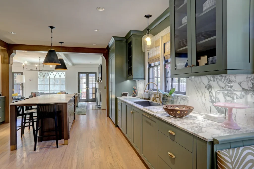Modern kitchen with green cabinets, marble countertops, a large island with seating, pendant lights, and wood flooring. Natural light enters through windows and a glass-panel door.