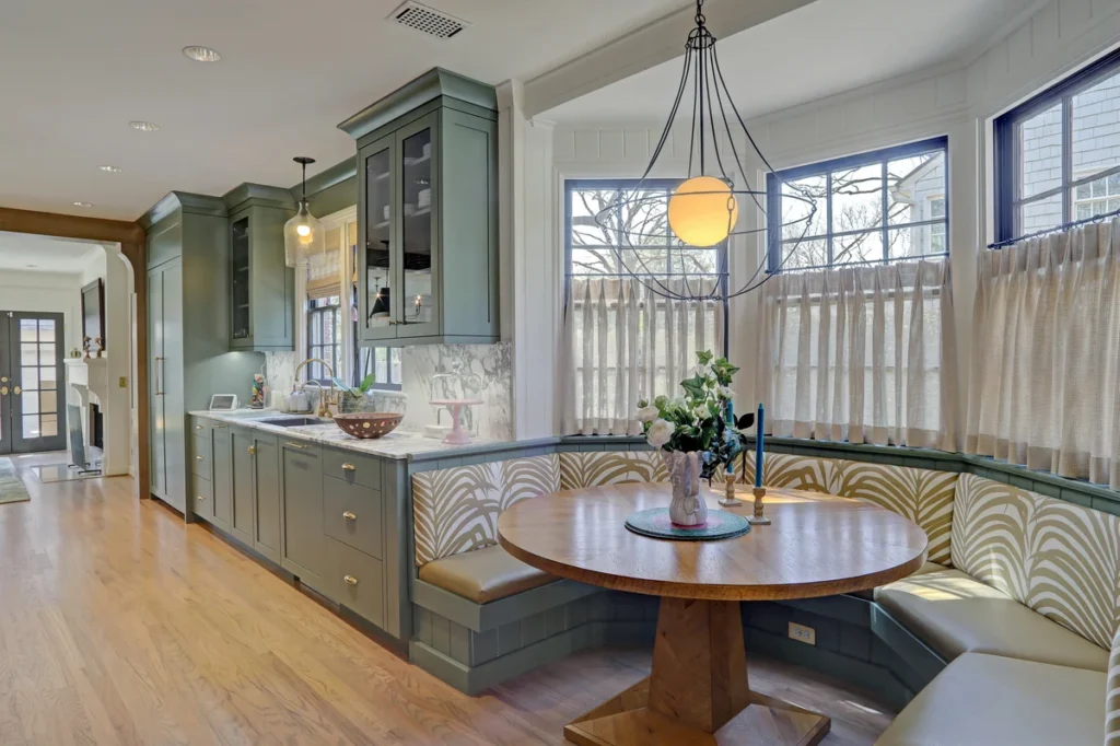 A kitchen with green cabinets, light wood flooring, a round wooden table with built-in bench seating, and large windows with half curtains.