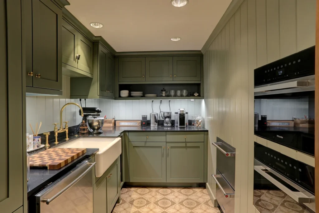 A compact kitchen with green cabinets, built-in ovens, a farmhouse sink, various small appliances, and patterned tile flooring.