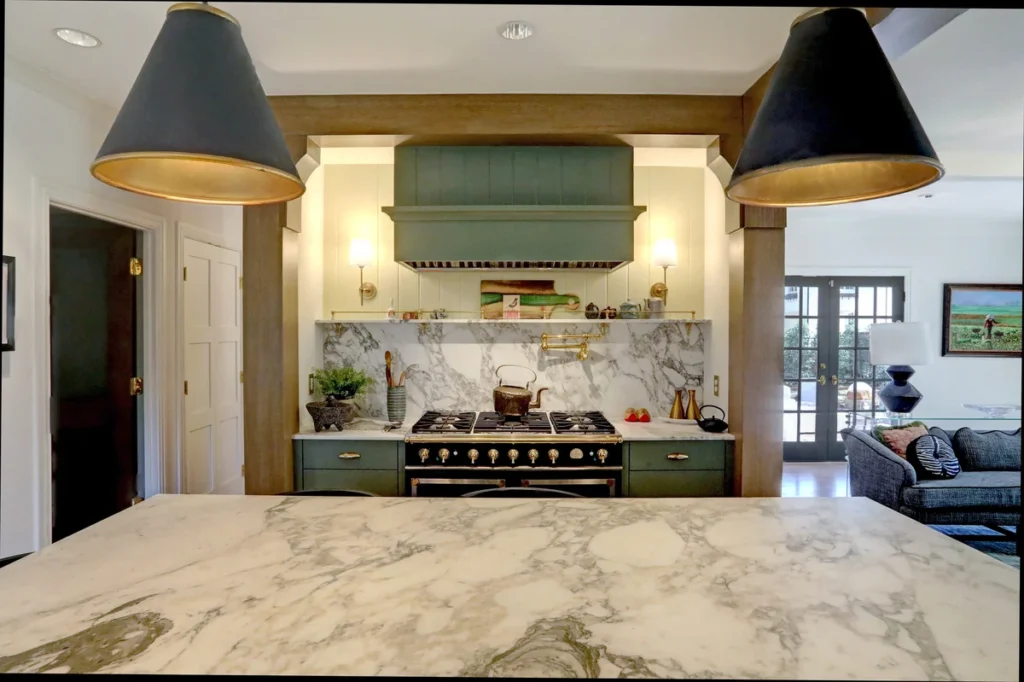 Modern kitchen with green cabinetry, marble countertops, large black pendant lights, a gas stove, and decorative backsplash; living room visible in the background.