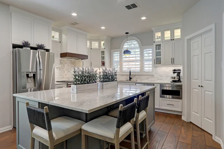 Modern kitchen with white cabinets, stainless steel appliances, a large island with four chairs, and decorative plants on the countertop.