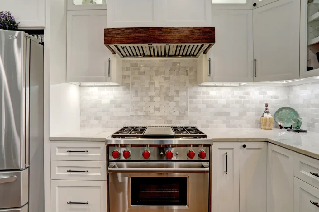 Modern kitchen with a stainless steel gas stove, oven, and a wood-accented range hood, surrounded by white cabinets and a tiled backsplash.