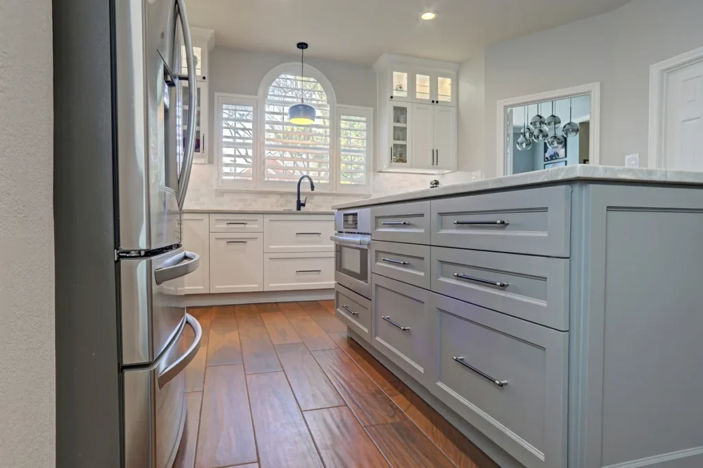 Modern kitchen with stainless steel refrigerator, gray and white cabinetry, large island, wooden floors, and arched window with shutters.
