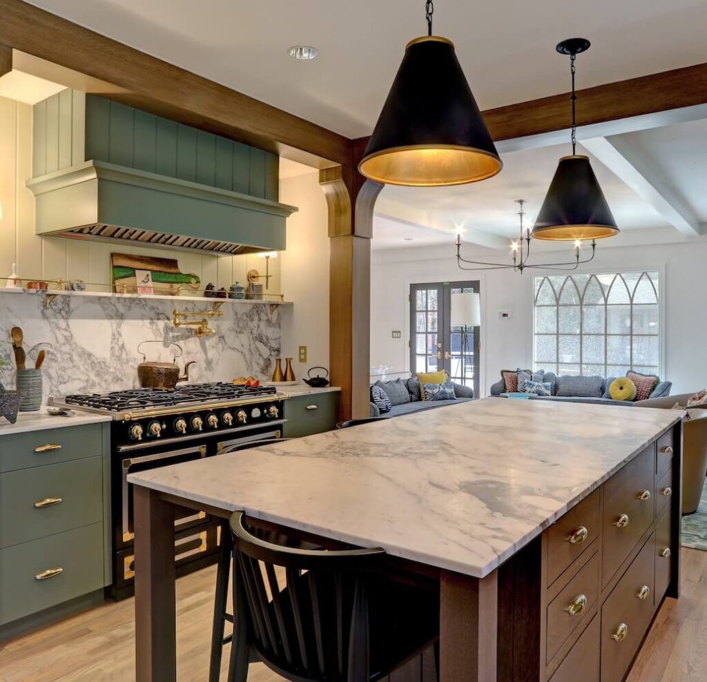 Modern kitchen with green cabinets, marble countertops, a large island, black and gold stove, hanging black pendant lights, and an open view into a living area with large arched windows.