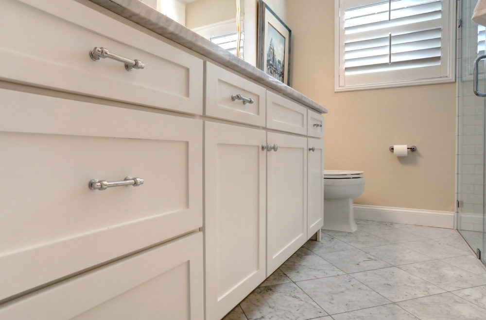White bathroom cabinets with silver handles are set against a marble tile floor; a toilet, toilet paper holder, and window with shutters are visible in the background.