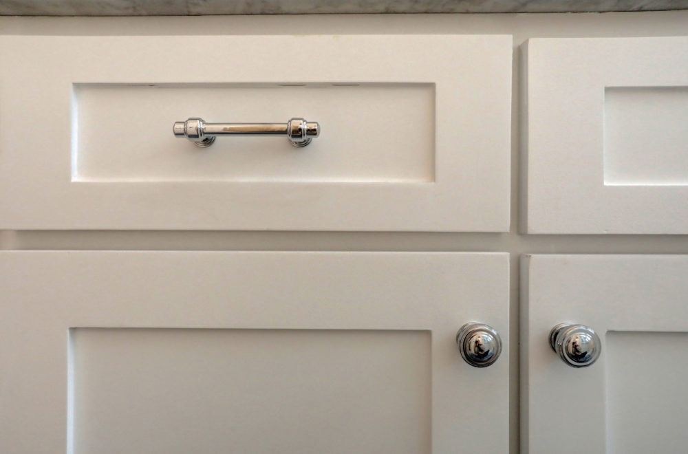Close-up of white cabinet doors and a drawer with silver knobs and a silver handle, set against a light-colored countertop.