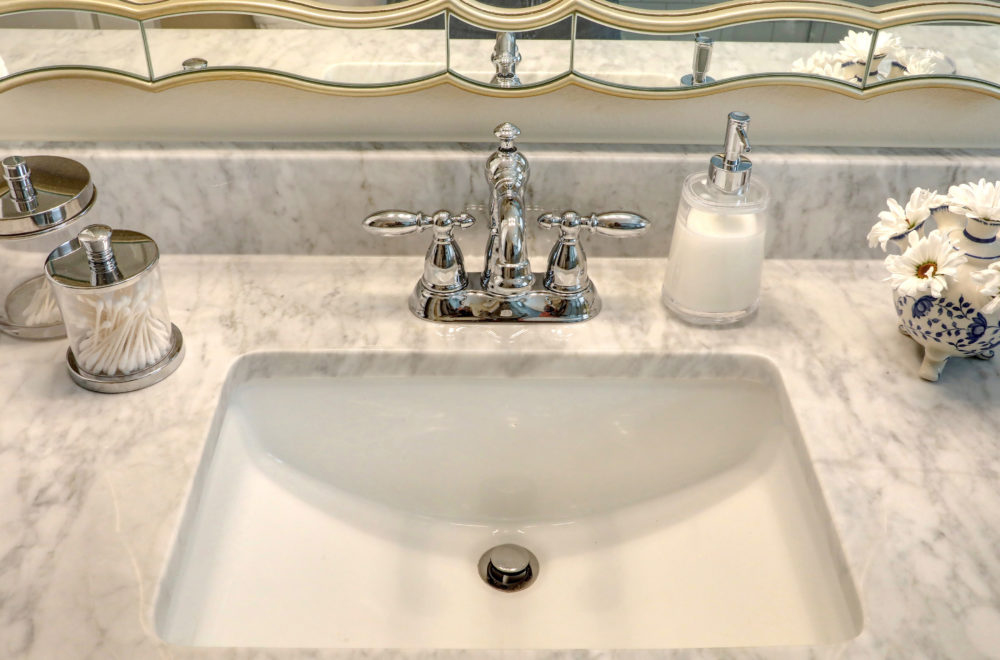 A bathroom sink with a marble countertop, chrome faucet, a soap dispenser, a cotton swab container, and decorative items beside a mirror.