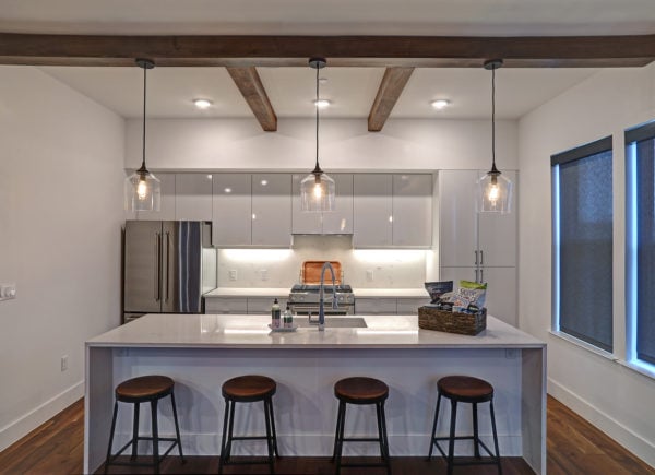 Modern kitchen with white cabinets, a marble island with sink, four wooden stools, pendant lights, and exposed wooden ceiling beams.