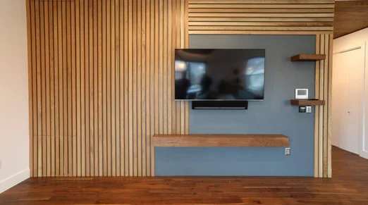 Magnolia-Loft-05-1000x660 A modern living room wall with vertical wooden slats, a mounted flat-screen TV, floating shelves, and a wooden bench against a gray accent panel.