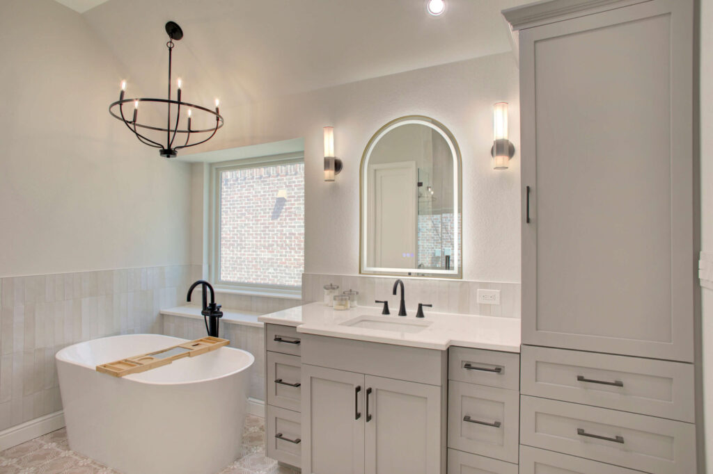 Modern bathroom with a freestanding tub, black fixtures, a large window, gray cabinetry, and a vanity with an arched mirror and wall sconces.
