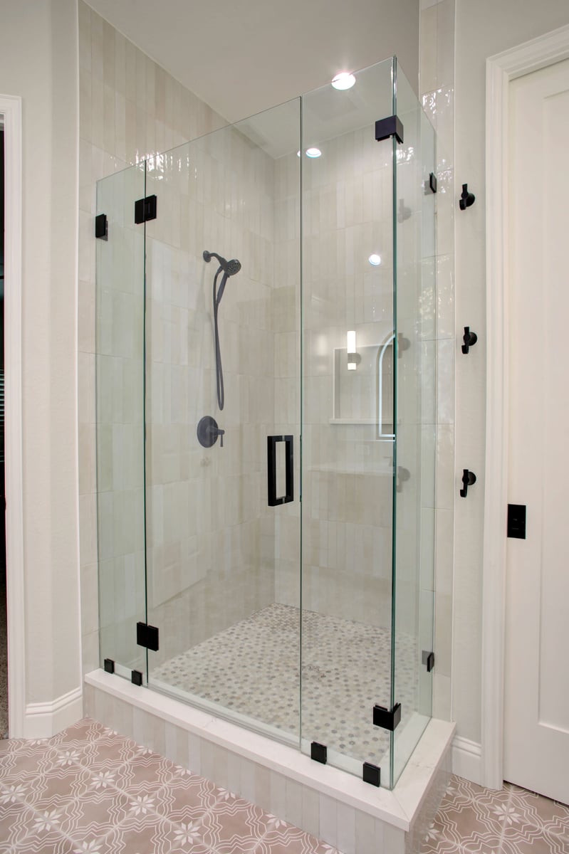 Modern glass-enclosed shower with black hardware, beige tiled walls, built-in niche, and patterned tile floor inside and outside the shower area.