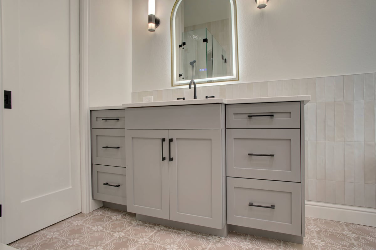 Light gray bathroom vanity with black handles, white countertop, arched mirror, wall sconces, and patterned tile floor. White wall tiles are partially visible behind the vanity.
