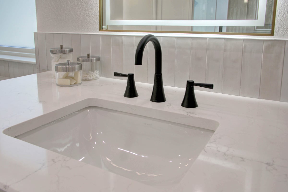 A modern bathroom sink with a white marble countertop, black faucet and handles, and clear jars holding cotton pads and swabs.