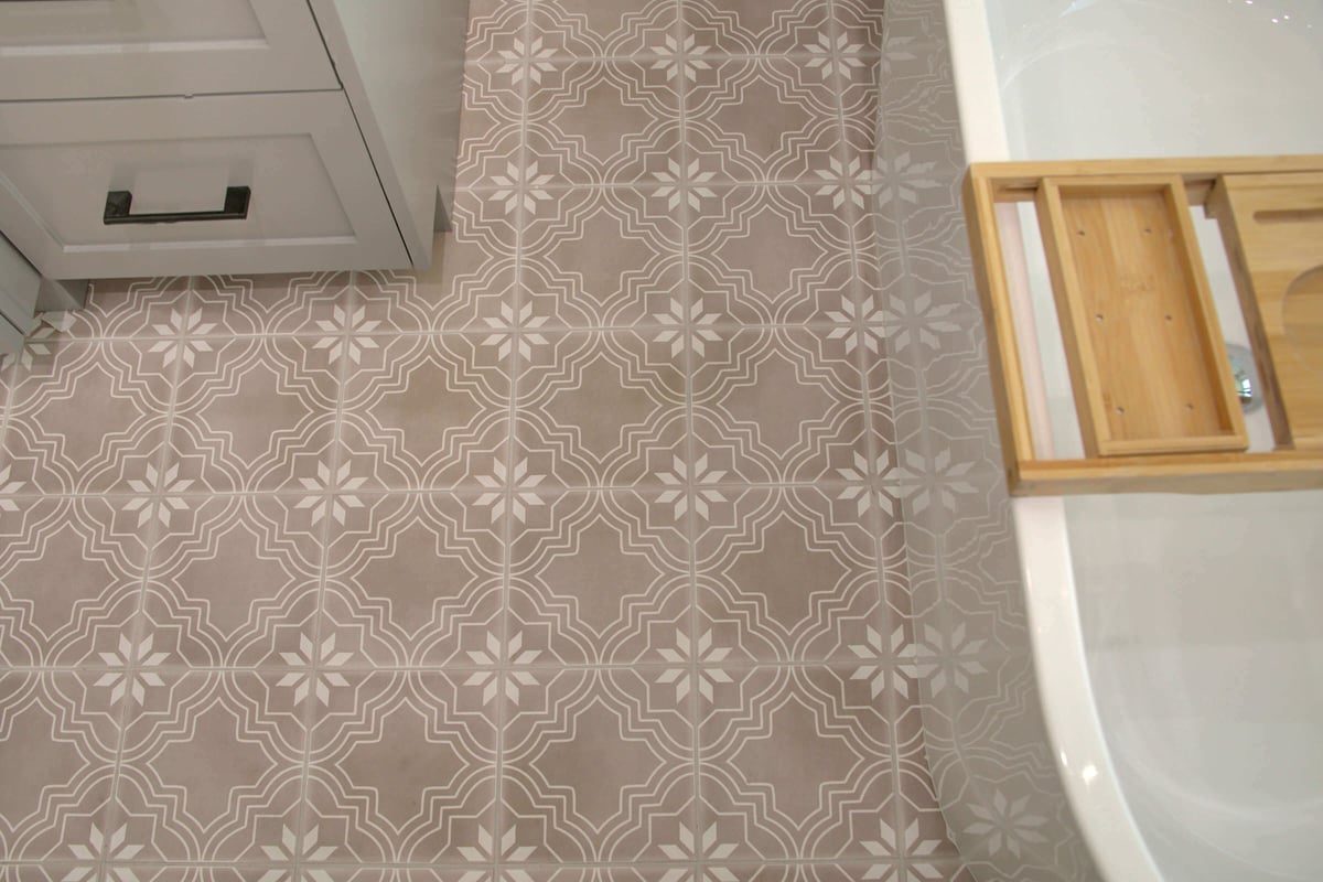 Gray tile floor with a white geometric and floral pattern next to a white cabinet and a bathtub with a wooden bath tray.